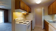 This single-floor space includes a compact kitchen designed for practicality and ease of use. The kitchen is equipped with a white electric coil-burner range and a matching white refrigerator, offering essential cooking and food storage functionality. Warm-toned wooden upper cabinets line the wall above the range, providing ample storage, while an under-cabinet range hood with built-in lighting serves both ventilation and task lighting needs. The walls are finished in a neutral lavender-gray tone, contributing to a calm, understated ambiance. Light tile flooring runs through the kitchen and continues into the hallway beyond, creating a cohesive flow throughout the space. A flush-mount ceiling light fixture provides overhead illumination. A doorway visible at the end of the kitchen corridor leads to an adjacent room, suggesting an efficient and connected layout. The textured ceiling adds a subtle architectural detail consistent with the straightforward, functional character of the overall space. This kitchen reflects a no-frills design focused on utility and ease of movement within a modest footprint.
