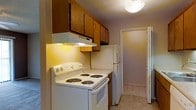 This single-floor space includes a compact kitchen designed for practicality and ease of use. The kitchen is equipped with a white electric coil-burner range and a matching white refrigerator, offering essential cooking and food storage functionality. Warm-toned wooden upper cabinets line the wall above the range, providing ample storage, while an under-cabinet range hood with built-in lighting serves both ventilation and task lighting needs. The walls are finished in a neutral lavender-gray tone, contributing to a calm, understated ambiance. Light tile flooring runs through the kitchen and continues into the hallway beyond, creating a cohesive flow throughout the space. A flush-mount ceiling light fixture provides overhead illumination. A doorway visible at the end of the kitchen corridor leads to an adjacent room, suggesting an efficient and connected layout. The textured ceiling adds a subtle architectural detail consistent with the straightforward, functional character of the overall space. This kitchen reflects a no-frills design focused on utility and ease of movement within a modest footprint.