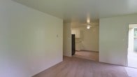 This single-floor residential space offers a straightforward, functional layout designed for comfortable everyday living. The main living area features neutral-toned wall-to-wall carpeting that extends throughout, creating a warm and cohesive feel across the space. Smooth, light-colored walls and standard ceilings contribute to a clean, understated aesthetic throughout.

The living area flows openly into the kitchen, which is visible from the main room and separated by a partial wall. The kitchen is equipped with white cabinetry providing ample storage, a black refrigerator, and a countertop workspace, offering a practical setup for meal preparation. A ceiling fan is centrally positioned overhead, providing both ventilation and a source of ambient lighting for the combined living and kitchen area.

The open sightlines between the living and kitchen areas enhance the sense of connectedness within the space, making it feel accessible and easy to navigate. The neutral color palette throughout — from the carpeting to the walls and cabinetry — creates a versatile backdrop that accommodates a variety of interior styles. Overall, this single-level space presents a practical and livable environment with a simple, unpretentious design well-suited for day-to-day use.