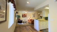 This single-floor residence offers a thoughtfully designed open-concept layout that promotes a natural flow between the main living areas. The living room is a comfortable and inviting space, featuring rich hardwood-style flooring, a ceiling fan with integrated lighting, and warm neutral wall tones that create a relaxed atmosphere. A cozy seating arrangement anchored by a plush area rug defines the space, while recessed lighting and a decorative lamp provide layered illumination throughout the day and evening.

The kitchen is positioned adjacent to the living area and separated by a peninsula counter with white cabinetry, offering both functionality and an open sightline to the rest of the home. The kitchen features ample cabinet storage, a built-in cooktop, and a bright, clean aesthetic with light-toned countertops. Decorative touches such as fresh flowers and fruit add a welcoming character to the space.

Natural light filters in through generously sized windows, enhancing the airy feel of the interior. The textured ceiling and clean architectural lines throughout contribute to a classic residential style. Overall, this single-floor layout offers a practical and comfortable living environment with a warm, cohesive design sensibility well-suited for everyday living.