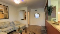 This single-floor residence showcases a thoughtfully designed interior that blends classic architectural elements with a warm, inviting ambiance. The living area serves as the centerpiece of the space, featuring hardwood-style flooring that extends throughout, creating a cohesive and polished look underfoot. Crown molding lines the upper edges of the walls, adding a refined finishing touch to the interior.

A defining architectural feature is the prominent arched entryway that frames the transition between the living area and an adjacent room, lending an elegant, classic character to the layout. A secondary arched niche adds further visual interest and provides a display space. Complementing these design elements is a decorative circular window mounted on an accent wall, serving as a unique focal point that draws the eye and introduces natural light in an unexpected way.

The living room is furnished with a light-toned sofa and chaise arrangement, paired with a round coffee table and decorative accessories that contribute to the space's welcoming feel. A ceiling fan with a light fixture provides both air circulation and ambient illumination. The neutral, warm wall tones throughout create a calm and comfortable atmosphere suited for everyday living.