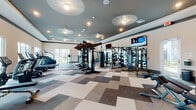 This single-level fitness center is thoughtfully designed to accommodate a wide range of workout styles and fitness goals. The space is divided into two primary zones: a main gym floor and a dedicated studio room, each with distinct flooring and finishes that define their purpose.

The main gym area features a geometric-patterned carpet tile floor in neutral tones of gray, beige, and brown. It is outfitted with an extensive selection of TRUE-brand cardio equipment, including treadmills, ellipticals, and stationary bikes, positioned along large windows that provide natural light and views of the surrounding outdoor area. A robust strength training section includes a multi-functional cable and power rack system, free weights, dumbbells, kettlebells, medicine balls, and a heavy punching bag. Wall-mounted flat-screen TVs are positioned for easy viewing. Ceiling-mounted circular light fixtures complement the recessed lighting throughout, and a water fountain station is conveniently accessible near an exterior door.

The adjoining studio room features warm hardwood-style flooring, tray ceilings with recessed lighting, full-length wall mirrors, and adjustable benches, making it well-suited for stretching, yoga, or functional training. Glass double doors connect the studio to an outdoor walkway, allowing ample natural light to fill the space.