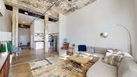 Located at 2450 Roosevelt Ave in San Antonio, TX, this one-bedroom, one-bathroom loft delivers a striking industrial aesthetic defined by soaring ceilings, exposed concrete columns, and raw, weathered ceiling details that pay homage to the building's architectural heritage. Polished concrete floors run throughout, anchoring the open-concept layout with a cohesive, modern edge.

The spacious living area is bathed in natural light from large windows fitted with blinds, creating a bright and airy atmosphere. A wall-mounted mini-split unit provides climate control, while the open floor plan allows for flexible furniture arrangements. The kitchen sits adjacent to the living space and features stainless steel appliances — including a full-size refrigerator, range, and dishwasher — paired with open metal shelving and white cabinetry topped with dark counters.

A hallway connects the living area to the private bedroom, which includes a closet and offers a cozy yet functional retreat. The bathroom features warm-toned flooring and clean white finishes. Throughout the unit, the interplay of white walls against aged concrete and exposed structural elements creates a compelling contrast that defines the loft's unique urban character.