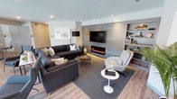 This single-floor space showcases a thoughtfully designed living area that blends modern aesthetics with functional comfort. The focal point of the room is a striking floor-to-ceiling feature wall clad in large-format grey tile, housing a sleek linear electric fireplace beneath a wall-mounted television. Flanking the feature wall, built-in open shelving provides both display and storage space, adding architectural interest while keeping the design cohesive. Light hardwood flooring runs throughout, contributing warmth and continuity to the space. The living area is generously proportioned, comfortably accommodating a large dark sectional sofa paired with a light accent chair and a round glass coffee table, creating a balanced and inviting seating arrangement. Recessed lighting in the white ceiling ensures even illumination throughout, while a floor lamp adds a layered lighting element. Neutral tones of grey, white, and warm wood dominate the palette, lending the space a clean, contemporary feel. A framed artwork piece on the adjacent wall adds a finishing touch to this well-appointed, modern interior.