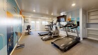 Located at 450 N Armando St in Anaheim, California, this single-floor fitness center is a well-appointed exercise facility designed to support an active lifestyle. The main gym area serves as the heart of the space, equipped with multiple treadmills, elliptical machines, and stationary bikes arranged on protective floor mats over carpeted flooring. A large wall-mounted mirror spans one side of the room, creating a sense of expanded space while allowing users to monitor their form, and a flat-screen TV is mounted above for entertainment during workouts. Recessed lighting illuminates the room evenly throughout. A standout feature is the bold, colorful mural adorning one wall, depicting palm trees and tropical motifs in vivid shades of teal, orange, and navy — adding a lively, energetic character to the environment. French doors connect the gym to an outdoor patio, which offers a covered concrete area with seating and open sky views, ideal for stretching or a post-workout cooldown. Additional rooms include a dedicated entrance, a utility room, and a closet with built-in shelving for storage. The overall layout is efficient and purposeful, making excellent use of the available footprint to deliver a comprehensive, community-oriented fitness experience.