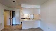 This single-floor space offers a practical and efficient layout designed to maximize functionality. The kitchen serves as a central feature, defined by a peninsula-style counter that seamlessly separates it from the adjacent living area. The cooktop is integrated directly into the counter surface, complemented by a sink and a full-sized refrigerator tucked neatly within the cabinetry. Expansive upper cabinets with a clean, flat-front design provide generous storage, while under-cabinet lighting adds both utility and a subtle warmth to the workspace. The kitchen's tile flooring transitions naturally into the carpeted living area, creating a clear yet open delineation between spaces. A dedicated laundry area is conveniently situated just off the kitchen, housing a full-sized washer and offering additional built-in shelving for storage. Throughout the space, a consistent neutral color palette of whites and soft beiges lends a bright, airy feel. The textured ceiling adds a classic architectural detail, and the overall layout reflects a straightforward, well-organized design that prioritizes ease of living.