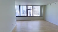 This single-floor space is defined by its striking floor-to-ceiling windows that span the width of the main living area, offering unobstructed, elevated views of a dense urban skyline. The large window panels, framed in dark trim, allow abundant natural light to flood the interior throughout the day, creating a bright and airy atmosphere. The main living area features smooth, light-toned hardwood flooring that enhances the sense of openness and reflects the natural light from the expansive windows. The walls are finished in a clean, neutral white, contributing to the minimalist aesthetic of the space. The open layout provides a versatile canvas, allowing for flexible furniture arrangements. The high-rise positioning of the unit places it well above neighboring structures, ensuring privacy while maximizing panoramic city views. The overall ambiance is modern and clean, with an emphasis on natural light and urban connectivity. Practical details include floor-level ventilation registers integrated discreetly into the baseboard area, maintaining the streamlined appearance of the interior.