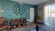 Located at 6300 Pewter Ave in North Chesterfield, Virginia, this single-floor home offers a well-organized and functional layout across several distinct living spaces. The living room serves as the heart of the home, featuring plush carpeting, a bold teal accent wall, and a large sliding glass door that fills the space with natural light. A ceiling-height decorative clock and a cozy seating arrangement contribute to the room's inviting atmosphere. Adjacent to the living area, the dining space accommodates a four-seat round table beneath a classic overhead light fixture, with direct access to the kitchen. The kitchen is outfitted with white cabinetry, white appliances including an electric range and refrigerator, and ample counter space, maintaining a clean and practical aesthetic. The primary bedroom is carpeted and features a ceiling fan, a closet with louvered doors, and a window with natural light. A second bedroom offers similar carpeted flooring, a window with curtains, and space for a desk, making it versatile for sleeping or working. The full bathroom includes a tub-shower combination. Throughout the home, neutral wall tones and textured ceilings provide a consistent, understated backdrop that complements each room's functionality.