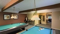 This single-floor entertainment space is designed with recreation and socializing in mind. The centerpiece of the room is a pair of full-size pool tables, each outfitted with vibrant blue felt and illuminated by classic pendant-style billiard lights suspended from chains above — providing both focused task lighting and a warm, inviting atmosphere. The ceiling features a textured finish with exposed dark wood beams that add a rustic, grounded character to the space. Walls are painted in a rich chocolate brown, creating a cozy and dynamic backdrop that contrasts nicely with the lighter accents throughout. A bar-height table with tall stools offers a casual seating area, ideal for spectators or between-game relaxation. An arched entryway leads to an adjoining area, adding architectural interest and a sense of flow. Decorative elements, including framed wall art, tall potted plants, and a charming oversized stuffed bear, lend personality and a playful spirit to the room. The carpeted flooring ties the space together and helps absorb sound, enhancing comfort during extended use. Overall, this game room delivers a well-appointed and lively setting suited for leisure and entertaining.