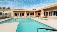 This single-story community amenity space is designed with outdoor living and recreation at its core. The centerpiece is a generously sized swimming pool with crystal-clear turquoise water, complemented by a separate spa/hot tub, both set within a spacious concrete pool deck that provides ample room for lounging. Orange-toned patio furniture and chaise lounges are arranged throughout the deck, offering comfortable spots to relax under the open sky. A covered outdoor seating area with large windows and French doors connects the interior clubhouse to the pool area, creating a seamless indoor-outdoor flow. The Mediterranean-inspired architecture, characterized by stucco exteriors and terra cotta tile rooflines, lends a warm, resort-like ambiance to the setting. A dedicated outdoor kitchen and barbecue area features a built-in multi-burner gas grill beneath a white pergola, paired with a dining table and chairs — perfect for casual gatherings. Surrounding landscaping includes desert-adapted plantings and palm trees, evoking a dry, sunny climate. The combination of the pool, spa, shaded pergola, and outdoor kitchen makes this amenity space a comprehensive and inviting destination for leisure and social activity.