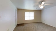 This single-story space features a welcoming main room finished with wall-to-wall neutral carpeting that lends a warm, comfortable feel underfoot. Near the entry door, the flooring transitions to durable hard surface material, providing a practical area for arriving and departing. A centrally positioned window allows natural light to fill the room, brightening the neutral-toned walls and contributing to an open, airy atmosphere. A ceiling fan with an integrated light fixture is mounted overhead, offering both lighting and airflow control year-round. The walls are finished in a soft, warm neutral tone that complements the light carpeting, while the textured ceiling adds a subtle visual detail. The entry door is equipped with a standard lock, and a light switch panel is conveniently positioned nearby. The overall layout is straightforward and functional, with clean lines and a simple design that provides a blank canvas adaptable to a variety of furnishing arrangements. The space maintains a modest, practical character throughout.