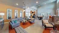 Located at 4090 Tradeway Dr in Leland, NC, this single-story community clubhouse offers an expansive, open-concept gathering space designed for both relaxation and socializing. The interior boasts soaring vaulted ceilings with high clerestory windows that flood the space with natural light, while polished concrete floors run seamlessly throughout, lending a modern, industrial character.

The main lounge area is anchored by a striking brick fireplace column with a mounted flat-screen TV above the hearth, surrounded by generous curved sectional seating, accent chairs, and warm orange area rugs that create an inviting atmosphere. Pendant globe lights hang overhead, complementing the two-tone warm orange and soft gray wall palette.

A dedicated billiards area with a full-size pool table sits adjacent to the lounge, adding a recreational dimension to the space. Beyond, a bar-style kitchen runs along the perimeter, featuring dark cabinetry, granite countertops, a utility sink, and a pass-through garage-style window that opens directly to an outdoor patio—ideal for entertaining. Exposed brick columns punctuate the interior, adding architectural texture and visual warmth throughout the clubhouse.