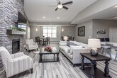 This single-floor space is designed with an open-concept layout that seamlessly connects multiple functional areas, creating a welcoming and sociable atmosphere throughout. Wide-plank wood-look tile flooring flows consistently across the entire space, tying each zone together with a warm, cohesive aesthetic.

The main living area serves as the heart of the space, anchored by a striking floor-to-ceiling stacked stone fireplace with a built-in hearth and mounted television above. Vaulted ceilings and recessed lighting enhance the sense of openness, while neutral earth-toned walls contribute to a calm, inviting ambiance. A ceiling fan adds both style and comfort to the central seating area.

Adjacent to the living area, a dedicated dining zone features a round pedestal table with high-back chairs, accented by large windows with plantation shutters that allow abundant natural light. Decorative wall sconces and framed artwork complete the refined atmosphere.

The kitchen is outfitted with dark espresso cabinetry, granite countertops, a classic white subway tile backsplash, and stainless steel appliances including a refrigerator, microwave, and dishwasher. A peninsula with bar-height seating bridges the kitchen to the main living area, making it ideal for casual gatherings.

Overall, the space offers a thoughtfully designed, multi-functional environment suited for relaxation, dining, and socializing.