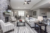 This single-floor space is designed with an open-concept layout that seamlessly connects multiple functional areas, creating a welcoming and sociable atmosphere throughout. Wide-plank wood-look tile flooring flows consistently across the entire space, tying each zone together with a warm, cohesive aesthetic.

The main living area serves as the heart of the space, anchored by a striking floor-to-ceiling stacked stone fireplace with a built-in hearth and mounted television above. Vaulted ceilings and recessed lighting enhance the sense of openness, while neutral earth-toned walls contribute to a calm, inviting ambiance. A ceiling fan adds both style and comfort to the central seating area.

Adjacent to the living area, a dedicated dining zone features a round pedestal table with high-back chairs, accented by large windows with plantation shutters that allow abundant natural light. Decorative wall sconces and framed artwork complete the refined atmosphere.

The kitchen is outfitted with dark espresso cabinetry, granite countertops, a classic white subway tile backsplash, and stainless steel appliances including a refrigerator, microwave, and dishwasher. A peninsula with bar-height seating bridges the kitchen to the main living area, making it ideal for casual gatherings.

Overall, the space offers a thoughtfully designed, multi-functional environment suited for relaxation, dining, and socializing.
