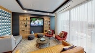 Located at 900 7th St SW in Washington, DC, this thoughtfully designed single-floor amenity space offers a refined blend of contemporary style and functional comfort. Expansive floor-to-ceiling windows wrapped in sheer white curtains flood the interior with natural light while framing sweeping views of the surrounding cityscape. The open layout is anchored by warm hardwood flooring that flows seamlessly throughout.

A well-appointed kitchen features teal cabinetry, a large wooden island, and ample counter space, making it ideal for both casual use and gatherings. Adjacent to the kitchen, a dining area showcases a round marble-top table beneath a striking lattice pendant light, surrounded by navy chairs — a sophisticated focal point complemented by lush potted greenery.

The lounge areas are equally inviting. A cozy seating nook with navy blue armchairs, warm pendant lighting, and a fiddle-leaf fig tree creates an intimate retreat bathed in natural light. A dedicated media lounge features built-in wooden shelving, a large wall-mounted screen, and eclectic seating including leather chairs and woven ottomans, set against a deep navy accent wall.

A reception or entry area includes double glass doors opening to an outdoor terrace with panoramic urban views. Dark-stained ceiling beam details and recessed lighting add architectural depth and polish throughout the entire space.