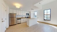 This single-floor unit showcases a thoughtfully designed open-concept layout that seamlessly connects the kitchen and living areas. The kitchen is a standout feature, outfitted with crisp white shaker-style cabinetry extending to the ceiling for generous storage, warm-toned countertops, and a full suite of stainless steel appliances including a refrigerator, microwave, stove, and dishwasher. A central island with matching countertop provides additional workspace and serves as a natural divider between the kitchen and the open living area. Light wood-look flooring runs throughout the main living space, lending warmth and continuity to the design. Large steel-framed windows flood the space with natural light and offer views of the surrounding area, enhancing the open, airy feel. Recessed lighting and a pendant fixture above the island provide layered illumination. The bedroom features neutral carpeting and a sliding double-door closet, offering a quiet and comfortable retreat. The bathroom is finished with light wood-tone tile flooring, a white vanity with warm countertop, a mirrored medicine cabinet, and clean, minimalist fixtures throughout.