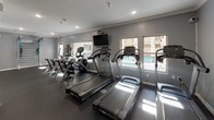 This fitness center offers a comprehensive workout environment thoughtfully designed to support a wide range of exercise routines. The space is finished with durable rubber flooring throughout, crown molding along the ceiling, and a neutral gray color palette that creates a clean, focused atmosphere. Recessed lighting keeps the space well-lit, while windows along one wall allow natural light to filter in and provide views of an adjacent outdoor pool area.

The cardio zone is well-stocked with multiple Cybex treadmills, elliptical machines, and stair climbers, each positioned to take advantage of the natural light and outward views. Wall-mounted televisions are available for entertainment during workouts. A dedicated strength and functional training area features a full dumbbell rack with a broad range of weights, a punching bag, a multi-use training station, and a medicine ball rack offering various weighted options. A large mirrored wall in the strength zone enhances the sense of space and assists with form checks during exercise.

An additional room adjacent to the main floor provides extra workout space, complete with motivational wall graphics and wood-look flooring, offering a versatile area for stretching, yoga, or additional training. Direct access to the outdoor pool area is provided through a glass-paneled door, seamlessly connecting indoor fitness with outdoor amenities.