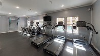 This fitness center offers a comprehensive workout environment thoughtfully designed to support a wide range of exercise routines. The space is finished with durable rubber flooring throughout, crown molding along the ceiling, and a neutral gray color palette that creates a clean, focused atmosphere. Recessed lighting keeps the space well-lit, while windows along one wall allow natural light to filter in and provide views of an adjacent outdoor pool area.

The cardio zone is well-stocked with multiple Cybex treadmills, elliptical machines, and stair climbers, each positioned to take advantage of the natural light and outward views. Wall-mounted televisions are available for entertainment during workouts. A dedicated strength and functional training area features a full dumbbell rack with a broad range of weights, a punching bag, a multi-use training station, and a medicine ball rack offering various weighted options. A large mirrored wall in the strength zone enhances the sense of space and assists with form checks during exercise.

An additional room adjacent to the main floor provides extra workout space, complete with motivational wall graphics and wood-look flooring, offering a versatile area for stretching, yoga, or additional training. Direct access to the outdoor pool area is provided through a glass-paneled door, seamlessly connecting indoor fitness with outdoor amenities.