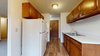 This single-floor unit is organized into a practical, functional layout that makes efficient use of its available space. The galley-style kitchen runs along two parallel walls and is outfitted with warm-toned wood cabinetry both above and below the countertops, providing ample storage. A white refrigerator and range occupy one side, while a double-basin sink with a standard faucet anchors the opposite counter. Wood-look plank flooring runs the length of the kitchen, leading toward a wood-paneled entry door at the far end. The living area transitions to neutral wall-to-wall carpeting and features clean white walls accented by wood-toned baseboard trim and a chair rail detail that runs along the perimeter. A sliding glass door opens to a balcony, allowing natural light into the space and offering exterior views, while an additional window and a wall-mounted air conditioning unit are also present. Baseboard heating runs beneath the windows throughout. The bathroom includes a full soaking tub with a tiled surround, a toilet, and a vanity with a light countertop, all set on mosaic tile flooring. Overall, the unit presents a straightforward, functional design with consistent finishes throughout.