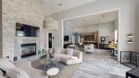 Located at 2300 Kathryn Ln in Plano, TX, this elegantly designed single-floor community space offers a refined and welcoming atmosphere. The open-concept layout seamlessly connects the living area and kitchen, creating a fluid and functional environment ideal for gathering.

The living area commands attention with its soaring ceilings, a dramatic floor-to-ceiling stacked stone fireplace with a wall-mounted TV, and a stunning tiered chandelier that anchors the room. Curved neutral sofas, a glass coffee table, and layered area rugs contribute to the upscale, contemporary aesthetic. Polished large-format tile flooring with a subtle wood-look pattern runs throughout, reflecting light and enhancing the sense of spaciousness.

A dedicated alcove features a grand piano set beneath a graceful arched bay window, offering serene views of the outdoor pool area and lush greenery — a standout architectural detail that fills the space with natural light.

The adjacent kitchen is well-appointed with granite countertops, gray cabinetry with glass-front upper doors, stainless steel fixtures, and warm wood-look flooring. A bright window above the counter adds natural light to the functional workspace.

Textured accent walls in deep gray tones and curated artwork complete the sophisticated, design-forward interior of this Plano community space.