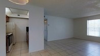 This single-floor space offers a straightforward, functional layout designed for comfortable everyday living. The main living area is open and generously proportioned, finished with light-toned ceramic tile flooring that runs throughout and contributes to a bright, airy feel. The walls are painted in a clean, neutral white, providing a versatile backdrop that maximizes the sense of space.

A partial dividing wall separates the living area from the kitchen zone, creating a subtle distinction between spaces while maintaining an open feel. The kitchen is positioned to one side and features dark cabinetry and standard appliances, including what appears to be a range, offering a practical cooking and prep area.

A bi-fold door is visible along one wall, likely concealing a closet or utility storage, adding to the unit's organizational functionality. The textured ceiling adds a subtle layer of detail to the otherwise streamlined interior, and overhead lighting provides adequate illumination throughout.

Overall, the space presents a clean, minimalist aesthetic with an emphasis on openness and practicality. The consistent tile flooring, neutral color palette, and efficient use of the floor plan make it a functional and adaptable living environment.