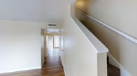 This two-story residence offers a thoughtfully designed layout that balances functionality and comfort. The interior is finished with rich hardwood flooring that runs throughout the main living areas, complementing the warm, neutral palette of the walls and ceilings. A central hallway connects the key rooms on the main level, drawing the eye toward a naturally lit space at the end anchored by vertical blinds that diffuse soft, ambient light throughout the day. The hallway provides a clean, unobstructed flow between rooms, reinforcing the home's practical and open feel. A staircase with a crisp white railing leads to the upper floor, adding a classic architectural element to the interior. Ceiling vents indicate a central HVAC system, ensuring climate control across both levels. The vaulted or angled ceiling detail visible near the staircase adds subtle architectural interest and a sense of vertical space. Overall, the home presents a clean, move-in-ready aesthetic characterized by consistent finishes, ample natural light, and a straightforward two-floor layout well suited for everyday living.