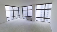 This single-floor space is defined by its striking wall-to-wall, floor-to-ceiling windows that dominate multiple sides of the room, flooding the interior with abundant natural light throughout the day. The expansive glazing frames views of surrounding high-rise buildings, offering a distinctly urban atmosphere with a sense of elevation and openness.

The interior features neutral-toned carpeting that extends across the full floor area, contributing to a clean and understated aesthetic. White walls and structural columns complement the light, airy feel of the space, while the open-plan layout provides significant flexibility for a variety of configurations or uses.

A wall-mounted HVAC unit is positioned centrally beneath one of the large window panels, ensuring climate control without compromising the streamlined design. The absence of partition walls creates an unobstructed flow throughout, making the space feel generously proportioned and adaptable.

Overall, the space presents a bright, minimalist environment with a strong urban character, well-suited for those seeking a versatile, light-filled setting in a high-rise setting.