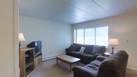 This single-floor residential space offers a practical and comfortable layout designed for everyday living. The living room serves as the central gathering area, featuring wall-to-wall carpeting that adds warmth underfoot and a neutral color palette throughout that creates a calm, understated ambiance. A large picture window dressed with vertical blinds allows generous natural light to fill the room, brightening the space and offering a view of the surrounding greenery outside. The living area comfortably accommodates a full seating arrangement, including a dark upholstered sofa and matching armchair positioned around a classic wooden coffee table — ideal for relaxing or entertaining. A media console along the adjacent wall provides dedicated space for a television and storage. The flat, paneled ceiling contributes to the clean, simple aesthetic of the interior. A baseboard heating unit runs along the exterior wall beneath the window, ensuring comfortable temperatures year-round. Overall, the space presents a no-frills, functional residential environment with a cozy atmosphere and straightforward design that prioritizes comfort and usability.