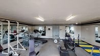 Located at 1570 Countryside Dr in Greenwood, Indiana, this fitness center is a dedicated, single-floor exercise facility designed to support a comprehensive workout routine. The space is appointed with a textured ceiling and a neutral color palette accented by bold horizontal striping along the walls, lending the room a structured, energetic atmosphere. Fluorescent lighting fixtures provide bright, even illumination throughout, complemented by natural light filtering in through latticed windows. Large wall-mounted mirrors span multiple sections of the room, visually expanding the space and allowing users to monitor their form during exercise. The flooring consists of durable carpeting in the main areas, with protective rubber matting—featuring a distinctive blue and yellow striped pattern—laid beneath select equipment for added safety. The strength training section is well-equipped with a barbell and weight bench, a squat/power rack, a multi-function cable machine, and a full dumbbell rack with a water cooler nearby. The cardio section offers a variety of machines including treadmills, elliptical trainers, and a stationary bike. A workout reference chart is posted on the wall near the exit for user convenience. The facility is secured with a keyed entry door, ensuring a private and controlled environment for all users.