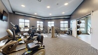 Located at 6620 Chestnut St in Lino Lakes, Minnesota, this fitness center offers a thoughtfully designed, single-floor exercise facility that caters to a wide range of workout needs. The space is divided into two distinct areas: a fully equipped cardio and strength training room, and a dedicated open-floor studio.

The main exercise room features textured carpet tile flooring and is outfitted with a comprehensive selection of equipment, including treadmills, recumbent bikes, elliptical machines, a NuStep cross trainer, and a multi-use strength machine. Multiple flat-screen televisions are mounted on the walls, and recessed lighting illuminates the space throughout. Rows of large windows allow abundant natural light to fill the room, creating an energizing atmosphere. Exposed ductwork along the upper walls adds an industrial design accent.

The adjoining studio area is finished in a calming blue-gray palette with smooth, light-toned hardwood-style flooring ideal for stretching, yoga, or group fitness. A full wall of mirrors maximizes the sense of space and supports movement-based workouts, while a mounted flat-screen television and wall-mounted stability balls and free weights provide additional functionality. Recessed and sconce lighting contribute to a clean, modern aesthetic. Together, the two areas form a versatile and well-appointed fitness facility.