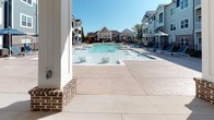 Located at 401 Neely Ferry Rd in Simpsonville, South Carolina, this single-story outdoor amenity space serves as a central gathering hub within a modern residential community. The centerpiece is a generously sized resort-style pool with in-water seating features, surrounded by an expansive concrete pool deck lined with lounge chairs and shaded by umbrellas—ideal for warm South Carolina days.

Adjacent to the pool, a covered pavilion provides a versatile outdoor living area designed for both relaxation and entertainment. The pavilion features a coffered white ceiling with multiple ceiling fans to ensure comfortable airflow. One end of the covered space includes a wall-mounted flat-screen television, a fireplace with brick surround, and stylish lounge seating with bold black-and-white accent pillows. A large white dining table with ample seating offers a dedicated space for outdoor meals and social gatherings.

The pavilion opens to the pool area through a wrought iron gated fence, blending seamless indoor-outdoor connectivity. The architectural detailing—including board-and-batten siding in muted blue-gray tones and exposed brick columns—complements the broader community's craftsman-inspired aesthetic, creating a cohesive and inviting atmosphere throughout.