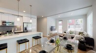 This well-designed single-floor residence offers a seamless open-concept layout that integrates the kitchen, dining, and living areas into one bright, cohesive space. The kitchen is a standout feature, equipped with stainless steel appliances, a dark tile backsplash, sleek white cabinetry extending to the ceiling, and a large waterfall-edge island with an integrated sink and bar seating illuminated by pendant lighting. Light wood-tone flooring flows throughout the main living areas, enhancing the airy, modern aesthetic. The dining area accommodates a glass-top table and benefits from generous natural light through large windows with outdoor views. The living room is anchored by expansive windows and a door providing direct access to an outdoor space, flooding the room with natural light. Two bedrooms offer carpeted flooring and ample natural light, with one featuring a walk-in closet. The two bathrooms are finished with warm wood-look flooring, floating gray vanities with white countertops, large mirrors, and clean white walls. One bathroom includes a full soaking tub and shower combination. Throughout, the residence maintains a clean, contemporary design with neutral tones, recessed lighting, and quality finishes that create a comfortable and stylish living environment.