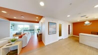 Located at 96 Coco Plum Dr in Marathon, FL, this single-floor space offers a thoughtfully designed combined living and dining area with a distinctly coastal Florida character. The living area is immediately eye-catching, featuring a bold orange accent ceiling that contrasts with crisp white walls and light wood-tone flooring throughout. Comfortable seating, including a spacious grey sofa accented with coral and neutral throw pillows, is arranged around a large flat-screen television mounted against a softly patterned blue-green accent wall. A wooden media console and open shelving add functional storage while maintaining the relaxed, beachy aesthetic. Large windows along the perimeter flood the space with natural light and provide views of the lush surrounding landscape. The dining area features a round white table with aqua wire chairs, contributing a playful, tropical feel. Glass-paneled interior walls and doors in the dining zone create visual openness and connectivity between areas. A small service counter with cabinetry is tucked nearby, adding practical utility. The overall ambiance is bright, welcoming, and distinctly Keys-inspired, blending vibrant color with casual, airy design elements suited to the warm South Florida environment.