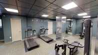 This fitness center is a dedicated exercise space designed to meet a variety of workout needs. The room features teal-blue painted walls that create a calm, focused atmosphere, complemented by a drop ceiling with recessed panel lighting that ensures bright, even illumination throughout the space. The flooring consists of patterned carpet tiles that provide cushioning and stability underfoot. Two treadmills with integrated digital displays are positioned along one wall, each equipped with touchscreen consoles for workout tracking and entertainment. A strength training machine is also present, rounding out the cardio and resistance options available. A distinctive diamond-plate metal column serves as a structural and decorative focal point near the center of the room, adding an industrial design element. Floor-to-ceiling mirrors line one wall, visually expanding the space and allowing users to monitor their form during exercise. A private door provides dedicated access to the facility. The combination of modern equipment, reflective surfaces, and purposeful layout makes this fitness center a practical and well-appointed amenity for regular use.