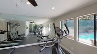 The fitness room at 924 Encinitas Blvd in Encinitas offers a practical and comfortable environment for exercise. The space is outfitted with a variety of cardio machines, including treadmills, elliptical trainers, and a stationary bike, as well as a multi-station cable weight machine to support a full range of strength training activities. Dark rubberized flooring provides a durable, slip-resistant surface throughout the room, while ceiling fans with integrated lighting ensure consistent air circulation and a well-lit atmosphere. Full-length mirrors line the walls, visually expanding the space and allowing users to monitor their form during workouts. Recessed lighting supplements the natural light that enters through multiple windows along the perimeter. A standout feature of this fitness room is its direct connection to the outdoor pool and patio area via a glass-panel door, offering views of the swimming pool and alfresco seating with umbrella-shaded tables. The neutral gray wall tones and clean white trim contribute to a tidy, focused environment. Crown molding adds a polished architectural detail to the room's interior. Overall, the fitness room delivers a functional and well-maintained space suited for a wide range of exercise routines.
