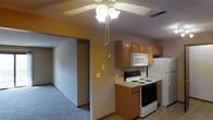 This single-floor residential space offers a practical and functional layout designed for comfortable everyday living. The kitchen is equipped with medium-toned wood cabinetry providing ample storage, light-colored countertops, and a full suite of white appliances including an electric range, over-the-range microwave, and refrigerator. The kitchen flows openly into the adjacent living area, creating a connected and accessible feel throughout. A ceiling fan with a light fixture is centrally positioned, offering both overhead lighting and air circulation. The living area features wall-to-wall carpet, contributing to a warm and quiet atmosphere. Neutral wall tones are used throughout, lending the space a clean, understated aesthetic. A textured ceiling adds subtle visual detail. The open passageway between the kitchen and the carpeted living space allows for easy movement and a sense of continuity across the floor plan. Overall, this space presents a straightforward, move-in-ready layout well-suited for those seeking a low-maintenance, single-level living environment.