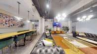 This single-floor amenity space blends industrial architecture with contemporary design to create a vibrant, multi-functional lounge environment. Polished concrete floors and exposed ceilings with visible ductwork and pendant lighting establish a raw, loft-like aesthetic throughout, while concrete structural columns reinforce the industrial character of the space.

The main lounge area serves as the social centerpiece, anchored by a tufted leather Chesterfield sofa, accent chairs with gold-tone frames, and a bold mustard area rug. A decorative lattice-patterned feature wall provides a striking backdrop for the wall-mounted flat-screen television, flanked by lush tropical plants that add natural warmth. A sculptural globe chandelier overhead adds a refined touch.

Adjacent to the lounge, a communal dining and bar-height seating area features a long wooden surface with green upholstered stools, ideal for collaborative gathering or casual dining. Built-in shelving and curated décor accents complete this zone.

The game and recreation area is a standout feature, highlighted by a full living green wall housing a mounted dartboard, and a high-top bistro table positioned before floor-to-ceiling steel-framed windows. These expansive windows flood the space with natural light and offer direct views to an outdoor courtyard area. The overall ambiance is energetic yet inviting, combining bold design choices with comfortable, communal-focused functionality.