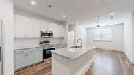 This well-appointed single-floor residence showcases a clean, contemporary design with a bright, neutral palette throughout. The heart of the home is an open-concept kitchen that flows seamlessly into the living area. The kitchen features a large central island topped with granite countertops and an undermount sink, complemented by two-tone cabinetry — crisp white upper cabinets paired with sage-green lower cabinets — set against a classic subway tile backsplash. Stainless steel appliances, including a range, microwave, refrigerator, and dishwasher, complete the modern look. A dedicated in-unit laundry area is conveniently tucked nearby. The open living space is finished with warm hardwood-style flooring and benefits from natural light through a large window, with a ceiling fan overhead adding comfort. The bedrooms are carpeted for warmth and comfort, each featuring ceiling fans and windows with blinds. The primary bedroom connects to a well-designed en-suite bathroom with a double granite-topped vanity, sage-green cabinetry, and hardwood-style flooring. A generously sized walk-in closet with built-in shelving and hanging rods provides ample storage. The overall ambiance is fresh, modern, and move-in ready.