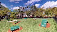 This outdoor dog park offers a welcoming and functional space designed with pets in mind. The grounds are covered in a well-maintained stretch of lush green grass, providing a comfortable surface for dogs to run, play, and exercise freely. The park is outfitted with a variety of green agility and enrichment equipment, including ramps, tunnels, elevated platforms, and balance features, giving dogs of all sizes and skill levels the opportunity to engage in stimulating physical activity. Red mulch borders accent the equipment areas, creating clear visual distinction and adding a neat, organized aesthetic to the layout. The space is fully enclosed by a secure black iron perimeter fence, ensuring safety for pets and their owners alike. Lush red-leafed hedges line the fence interior, adding a vibrant splash of color, while a generous canopy of mature palm trees and broad shade trees forms a natural backdrop, offering relief from the sun. The open sky above, with natural light flooding the area, enhances the inviting, outdoor ambiance. This park provides a clean, thoughtfully designed environment that encourages active use and socialization for dogs and their owners in a secure, green setting.