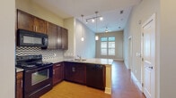 This single-floor residence offers a thoughtfully designed open-concept layout that seamlessly connects the kitchen and living areas. The kitchen is a standout feature, showcasing rich dark wood cabinetry, granite countertops, a mosaic tile backsplash, and a full suite of black appliances including a range, built-in microwave, dishwasher, and refrigerator. Pendant lighting and a track lighting system illuminate the space, while a peninsula countertop provides a natural divider between the kitchen and the adjoining living area. Warm hardwood-style flooring extends throughout the main living spaces, adding continuity and a welcoming ambiance. The living area benefits from elevated ceilings, a ceiling fan, and large windows that draw in generous natural light and views of the surrounding greenery. The bedrooms continue the bright, airy feel — one features hardwood-style flooring and multiple windows, while another is finished with plush carpeting and tall windows fitted with blinds. The bathroom is well-equipped with dark wood vanity cabinetry, a granite countertop with dual sinks, a large mirror, tiled tub/shower combination, and clean tile flooring. Throughout the residence, neutral wall tones and consistent finishes create a cohesive and inviting atmosphere.