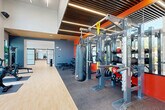 Located at 5851 Columbia Pike in Falls Church, VA, this professionally designed fitness center offers a comprehensive and contemporary workout environment across a single expansive floor. The space is defined by soaring ceilings, floor-to-ceiling windows along the exterior wall, and an abundance of natural light that floods the main exercise area, providing open views of the surrounding streetscape.

The main workout floor features light hardwood-style flooring transitioning to rubberized sport flooring in dedicated training zones. A full row of treadmills and elliptical machines lines the window wall, while a robust strength training section houses multi-station cable machines, Octagon power racks, battle ropes, weight plates, and heavy bags — all set against bold orange and red accent walls beneath a striking slatted wood and steel ceiling.

A separate functional training studio, accessible via stylish sliding barn-style glass doors, offers a clean, open space with mirrored walls, step platforms, yoga mats, and a large picture window overlooking the street. An additional flex room provides space for stretching, mat work, and small-group activities. The overall design balances energetic, motivating aesthetics with a clean, modern ambiance suited for a full range of fitness activities.