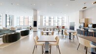 Located at 915 Division St in Nashville's dynamic Gulch neighborhood, this single-floor amenity space offers a generously sized, thoughtfully designed environment that blends communal living with professional functionality. The open-plan layout is anchored by soaring ceilings and an abundance of natural light streaming through expansive floor-to-ceiling windows, framing views of the surrounding Nashville skyline.

The main lounge area features distinct seating zones furnished with sculptural, rounded sofas and accent chairs in a curated palette of cream, burnt orange, and forest green, creating a vibrant yet cohesive aesthetic. Polished concrete-style flooring flows throughout, lending the space a clean, contemporary feel.

A café-style dining area provides numerous tables and chairs arranged for both casual dining and collaborative gathering, complemented by a service counter and display screens. Curated artwork adorns the walls, adding cultural character to the environment.

Private, glass-enclosed work suites line one side of the floor, each equipped with a dedicated desk, wall-mounted display, and accent seating — ideal for focused work or small meetings. Perforated metal and black steel framing define the partitions, introducing an industrial design element. Additional semi-private lounge nooks offer flexible spaces for quieter retreat within the broader community floor.