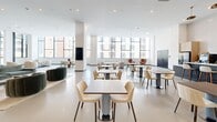 Located at 915 Division St in Nashville's dynamic Gulch neighborhood, this single-floor amenity space offers a generously sized, thoughtfully designed environment that blends communal living with professional functionality. The open-plan layout is anchored by soaring ceilings and an abundance of natural light streaming through expansive floor-to-ceiling windows, framing views of the surrounding Nashville skyline.

The main lounge area features distinct seating zones furnished with sculptural, rounded sofas and accent chairs in a curated palette of cream, burnt orange, and forest green, creating a vibrant yet cohesive aesthetic. Polished concrete-style flooring flows throughout, lending the space a clean, contemporary feel.

A café-style dining area provides numerous tables and chairs arranged for both casual dining and collaborative gathering, complemented by a service counter and display screens. Curated artwork adorns the walls, adding cultural character to the environment.

Private, glass-enclosed work suites line one side of the floor, each equipped with a dedicated desk, wall-mounted display, and accent seating — ideal for focused work or small meetings. Perforated metal and black steel framing define the partitions, introducing an industrial design element. Additional semi-private lounge nooks offer flexible spaces for quieter retreat within the broader community floor.