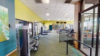 This spacious fitness center is designed to support a comprehensive workout experience, offering an extensive selection of equipment across a generously sized, open floor plan. The space is anchored by vibrant yellow and teal accent walls that lend an energetic ambiance throughout, complemented by a patterned wave-design carpet in green and gold tones that adds visual depth underfoot.

The cardio zone features multiple treadmills, elliptical trainers, stationary bikes — including a recumbent model — and rowing machines, all arranged to allow comfortable spacing between users. A dedicated strength training area houses a variety of cable machines, weight systems, and resistance equipment. Large floor-to-ceiling mirrors line one wall, visually expanding the space and providing users with form-checking visibility. Mounted flat-screen televisions offer entertainment during workouts.

Natural light filters in through multiple windows along the perimeter, keeping the environment bright and open. Large industrial-style fans provide additional air circulation throughout the floor. Colorful wall art and tropical plant accents contribute to a welcoming, resort-like atmosphere. A glass-walled corridor connects the fitness center to an open-air courtyard with stone tile flooring, offering a seamless transition between indoor and outdoor spaces. The drop ceiling with recessed panel lighting ensures consistent illumination across the entire area.