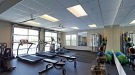 This single-floor fitness center is designed to support a comprehensive workout experience in a bright, open environment. The space is outfitted with a variety of cardio equipment, including treadmills and an elliptical machine, arranged to allow for easy movement between stations. A dedicated free weights area features a full rack of dumbbells, providing options for a wide range of strength training exercises. A multi-functional cable and strength machine anchors one side of the room, complemented by wall-mounted mirrors that enhance the sense of space and allow for proper form checks during workouts. The flooring consists of durable rubber matting, ideal for high-impact activities and equipment use. Overhead, a drop ceiling with recessed panel lighting ensures consistent, even illumination throughout. Natural light floods the space through multiple windows and a large garage-style roll-up door that opens to an outdoor area, creating an airy and inviting atmosphere. The overall layout is thoughtfully organized to accommodate both individual training sessions and small group workouts, making it a versatile and functional fitness facility.