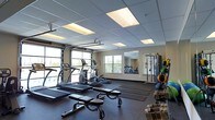 This single-floor fitness center is designed to support a comprehensive workout experience in a bright, open environment. The space is outfitted with a variety of cardio equipment, including treadmills and an elliptical machine, arranged to allow for easy movement between stations. A dedicated free weights area features a full rack of dumbbells, providing options for a wide range of strength training exercises. A multi-functional cable and strength machine anchors one side of the room, complemented by wall-mounted mirrors that enhance the sense of space and allow for proper form checks during workouts. The flooring consists of durable rubber matting, ideal for high-impact activities and equipment use. Overhead, a drop ceiling with recessed panel lighting ensures consistent, even illumination throughout. Natural light floods the space through multiple windows and a large garage-style roll-up door that opens to an outdoor area, creating an airy and inviting atmosphere. The overall layout is thoughtfully organized to accommodate both individual training sessions and small group workouts, making it a versatile and functional fitness facility.