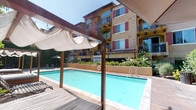 Located at 21535 Roscoe Blvd in the Canoga Park neighborhood of Los Angeles, this outdoor amenity area showcases a well-appointed resort-style pool complex set against the backdrop of a Mediterranean-inspired multi-story building. The architecture features warm golden stucco exteriors accented with bold red shutters and private balconies, lending the space a distinct Spanish Colonial character.

At the heart of the area lies a generously sized rectangular swimming pool with crystal-clear turquoise water, complemented by a separate spa for relaxation. The pool deck is finished in neutral concrete and natural wood decking, providing a clean and inviting surface for lounging. Elegant wood-framed cabana structures with canvas canopy shades offer shaded retreat areas, while additional umbrella-covered lounge chairs and a pergola-style gazebo provide further options for outdoor relaxation.

Overhead, strands of bistro string lights are draped across the space, creating a warm and festive ambiance during evening hours. The perimeter is thoughtfully landscaped with dense, lush hedging, flowering plants, and mature greenery that provides privacy and a verdant, tranquil atmosphere. A shaded patio seating area with a metal table and chairs offers a comfortable spot for casual dining or conversation. The overall environment is bright, open, and inviting, ideal for enjoying the Southern California sunshine.