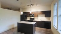 This single-floor space showcases a contemporary kitchen designed with a clean, modern aesthetic. Dark espresso-toned cabinetry lines the walls, offering generous upper and lower storage with sleek, understated hardware. The kitchen is anchored by a central island topped with a light countertop, providing additional workspace and a natural gathering point. Black appliances, including a refrigerator, range, microwave, and dishwasher, complement the dark cabinetry and contribute to the cohesive, uniform color palette. Light countertops along the perimeter contrast effectively against the darker elements, enhancing visual balance throughout the space. A stainless steel sink is positioned along the back wall, accompanied by ample counter space on either side. Overhead, a combination of hanging pendant lights and a track lighting system ensures thorough, well-distributed illumination across the workspace. Wood-look flooring runs throughout, adding warmth and texture that softens the otherwise bold, dark design choices. The open layout between the kitchen and the adjacent living area promotes a sense of spaciousness and ease of movement, making the space both functional and visually appealing.