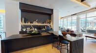 This expansive single-floor space is defined by a refined contemporary design, blending warm wood tones, dark matte finishes, and curated furnishings throughout its open layout. Light hardwood flooring flows seamlessly across the interior, contrasting with large-format tile in transitional zones and grounding each distinct area of the space.

A central bar and café station serves as a focal point, featuring dark stone countertops with dramatic veining, under-shelf accent lighting, and a boldly textured backsplash set against a deep charcoal accent wall. A wood-topped communal table with sleek black bar stools anchors the adjacent gathering area.

The lounge zones are thoughtfully arranged, offering a mix of intimate seating vignettes, built-in upholstered banquettes with bold yellow accents, and open social areas separated by elegant vertical slatted partitions. Warm cove lighting beneath wood-paneled ceiling insets adds depth and ambiance throughout.

Floor-to-ceiling curtain-wall glazing lines the perimeter, flooding the interior with natural light and framing sweeping city skyline views. Large sliding glass doors open directly onto a furnished outdoor terrace, extending the social space and offering additional seating with open-air city vistas.