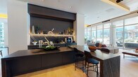 This expansive single-floor space is defined by a refined contemporary design, blending warm wood tones, dark matte finishes, and curated furnishings throughout its open layout. Light hardwood flooring flows seamlessly across the interior, contrasting with large-format tile in transitional zones and grounding each distinct area of the space.

A central bar and café station serves as a focal point, featuring dark stone countertops with dramatic veining, under-shelf accent lighting, and a boldly textured backsplash set against a deep charcoal accent wall. A wood-topped communal table with sleek black bar stools anchors the adjacent gathering area.

The lounge zones are thoughtfully arranged, offering a mix of intimate seating vignettes, built-in upholstered banquettes with bold yellow accents, and open social areas separated by elegant vertical slatted partitions. Warm cove lighting beneath wood-paneled ceiling insets adds depth and ambiance throughout.

Floor-to-ceiling curtain-wall glazing lines the perimeter, flooding the interior with natural light and framing sweeping city skyline views. Large sliding glass doors open directly onto a furnished outdoor terrace, extending the social space and offering additional seating with open-air city vistas.